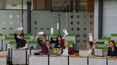 Studierende sitzen in einem Konferezsaal und halten Abstimmungskarten in die Höhe..