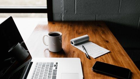 Foto von einem Tisch, auf dem ein Laptop, eine Tasse, ein Block mit Stift und ein Handy liegen