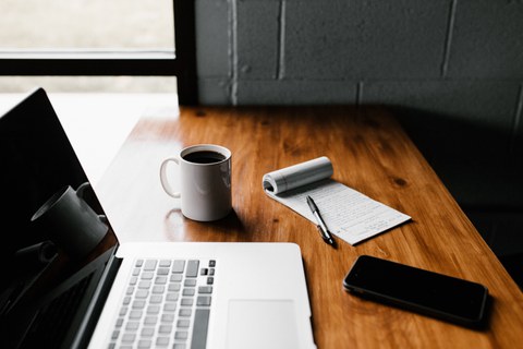 Foto von einem Tisch, auf dem ein Laptop, eine Tasse, ein Block mit Stift und ein Handy liegen