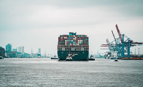 Ein großes, beladenes Containerschiff fährt durch einen Hafen. Auf der rechten Seite stehen Kräne 