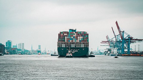 Ein großes, beladenes Containerschiff fährt durch einen Hafen. Auf der rechten Seite stehen Kräne 