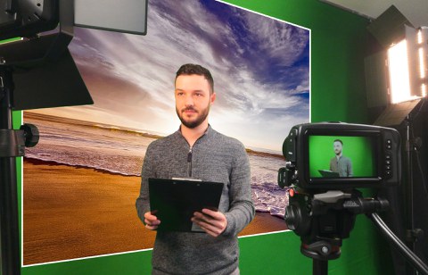 2 Scheinwerfer und eine Kamera sind auf eine Person vor einem Greenscreen gerichtet, wobei als Hintergrund ein digitales Bild von einem Sonnenuntergang am Meer eingefügt ist.