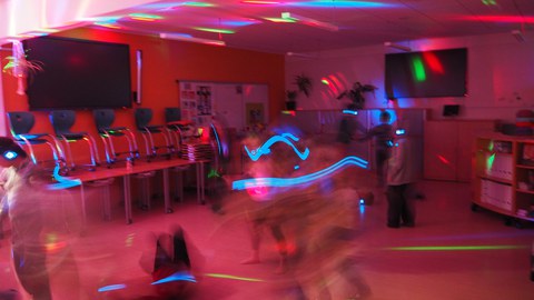 Klassenzimmer bunt beleuchtet bei Nacht, während Kinder tanzen. DIe Kinder sind durch die Bewegung sehr unscharf zu sehen. 