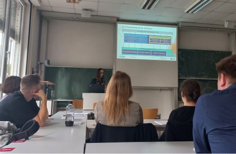 Foto: In einem Seminarraum steht eine Frau vor einer Tafel. Zu sehen sind zudem mehrere Zuhörer:innen in Rückenansicht.