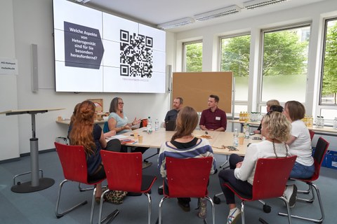 Eine Gruppe von Menschen sitzt um einen Tisch und bespricht sich zu einem Thema, das an der Powerpoint auf einer Leinwand rechts im Bild zu sehen ist.