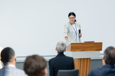 Das Foto zeigt eine Frau während eines Vortrags vor einer Seminargruppe. Sie steht hinter einem Rednerpult.