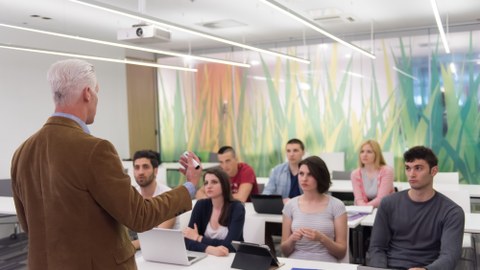 Foto eines Dozenten von hinten, der in einem Kurs zu 7 Studenten spricht und ihnen etwas erklärt.