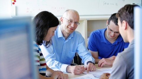 Bild von einem Dozenten und 3 Studierenden, die um einen Tisch sitzen und gemeinsam ein Paper durchsprechen. Im Hintergrund sind ein Holzregal und ein Whiteboard zu erkennen.