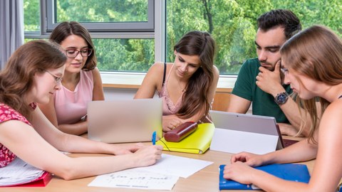 Foto von fünf Studenten, die in einer Workshoprunde zusammen am Laptop lernen.