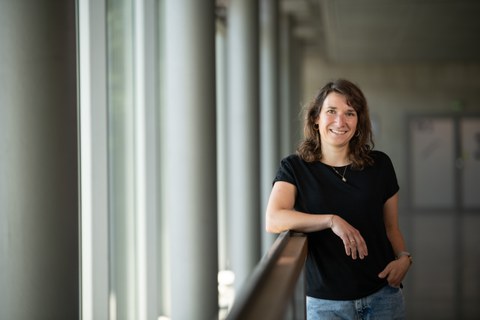 Auf dem Bild sieht man eine Frau, die an einem Geländer lehnt und in die Kamer lächelt.