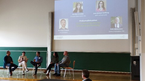 Einige Menschen sitzen in einem Saal vorne, dem Publikum zugewandt. Hinter ihnen an der Wand ist eine Powerpointpräsentation an einer Leinwand zu sehen.