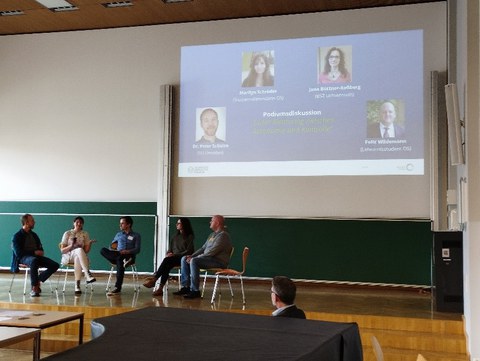 Einige Menschen sitzen in einem Saal vorne, dem Publikum zugewandt. Hinter ihnen an der Wand ist eine Powerpointpräsentation an einer Leinwand zu sehen.