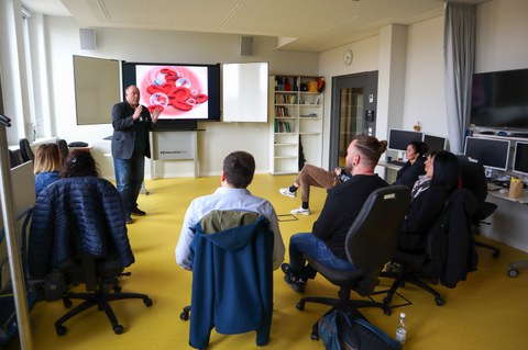 Auf dem Bild sind die Teilnehmenden des Workshops "Anforderungen an eine Befähigung auf dem Gebiet der didaktischen Medien aus der Sicht der Lehrenden" in einem Seminarraum zu sehen, die dem Dozenten zuhören.