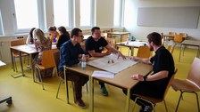 Das Bild zeigt Teilnehmende des Workshops "Gewalterfahrungen in der Rettungsdienstausbildung" diskutierend an einem Tisch in einem Seminarraum.