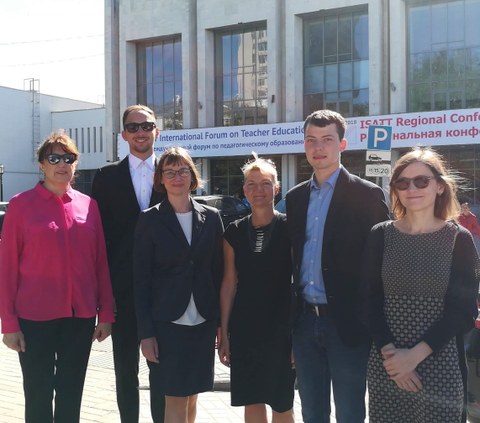 Foto einer Gruppe von 6 Personen, die vor einem Gebäude stehen. An dem Gebäude ist ein Banner mit Wörtern in Englisch und Russisch angebracht. "International Forum on Teacher Education" ist unter anderem zu lesen.