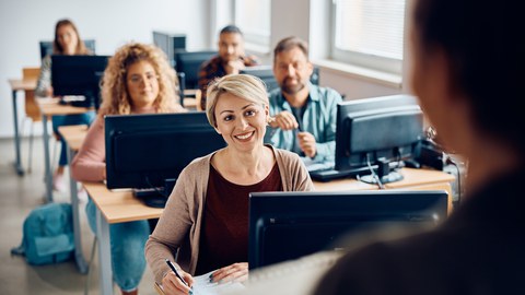 Das Foto zeigt einen Kursraum mit Computern auf den Tischen. An den Computern sitzen Personen und hören einem Kursleiter zu. 