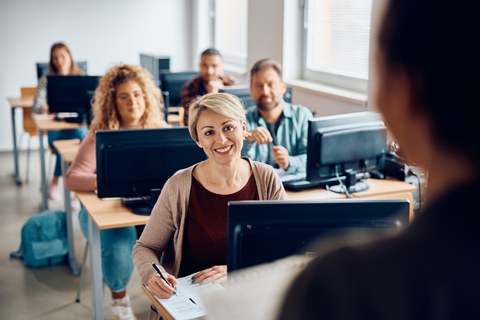 Das Foto zeigt einen Kursraum mit Computern auf den Tischen. An den Computern sitzen Personen und hören einem Kursleiter zu. 