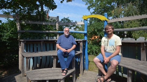 Foto: zwei Männer sitzen im Freien auf Holzbänken 