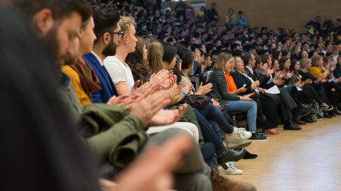 Foto eines großen Hörsaals, der voll belegt ist. Die sitzenden Personen klatschen.