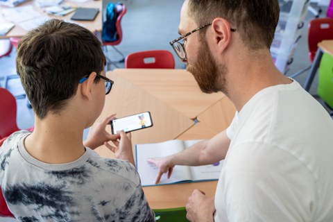 Das Foto zeigt einen Jungen und einen Mann von hinten. Der Mann erklärt dem Jungen etwas und zeigt dabei in ein Buch. Der Junge hält ein Smartphone in der Hand.