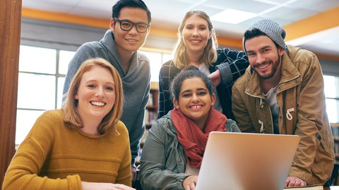 Foto einer Gruppe von 5 Studenten, die gemeinsam in der Bibliothek zusammen lernen und fröhlich in die Kamera lächeln.