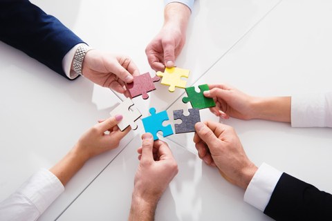 Sechs Hände verschiedener Personen halten sechs verschieden farbige Puzzleteile als Kreis in die Mitte. 