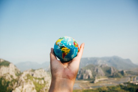 Foto mit einer Hand im Vordergrund, die einen kleinen Ball in Gestalt einer Weltkugel hochhält. Im Hintergrund ist eine Landschaft mit Bergen.