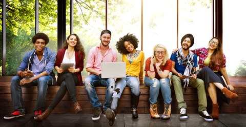 Bild einer Gruppe von sieben jungen Menschen, die nebeneinander auf einer Bank sitzen und in die Kamera lächeln