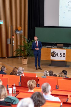 Moderator steht auf der Bühne und spricht zum Publikum.