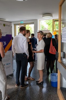Zwei Frauen und ein Mann unterhalten sich, während im Hintergrund buntes Treiben herrscht.