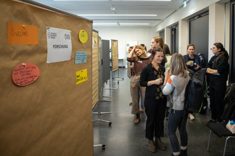 Das Foto zeigt eine Pinnwand mit Personen im Hintergrund, die sich unterhalten.