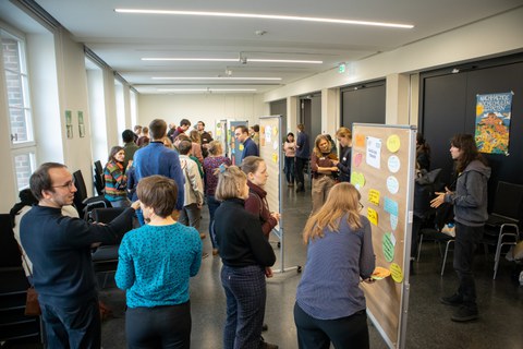 Das Foto zeigt einen raum gefüllt mit Menschen, die sich unterhalten und einer Reihe von Pinnwänden.