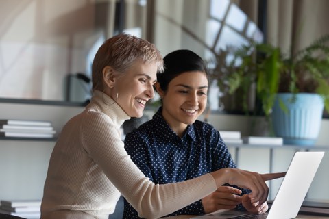 Zwei Frauen sitzen vor einem Laptop. Eine der Protagonistinnen zeigt etwas auf dem Bildschirm.