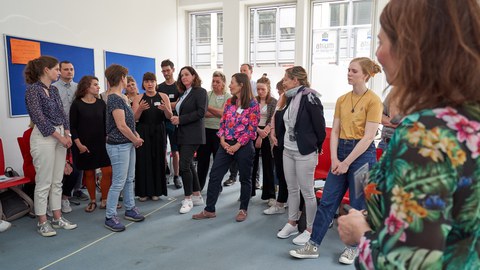 Eine Gruppe von Personen, die im Kreis stehen und einer Frau zuhören.