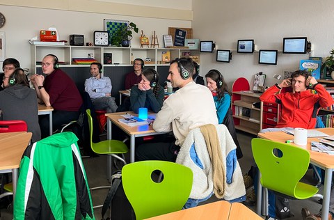 Auf dem Bild sind ca. zehn Erwachsene zu sehen, die mit Kopfhörern in einem inklusiven Klassenzimmer sitzen und einem Vortrag folgen. 