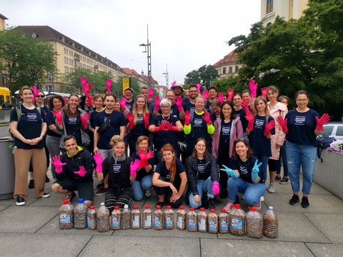 Eine große Gruppe von Personen, wo fast alle pinke Gummihandschuhe tragen. Vor der Gruppe sind viele Behälter zu sehen, die mit Zigarettenstummeln gefüllt sind.