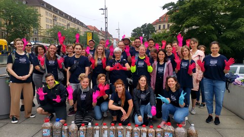 Eine große Gruppe von Personen, wo fast alle pinke Gummihandschuhe tragen. Vor der Gruppe sind viele Behälter zu sehen, die mit Zigarettenstummeln gefüllt sind.