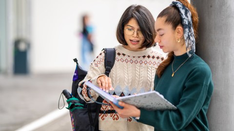 Foto zeigt, wie eine Studierende an einer Säule steht und ihre Notizen einer anderen Studierenden zeigt.