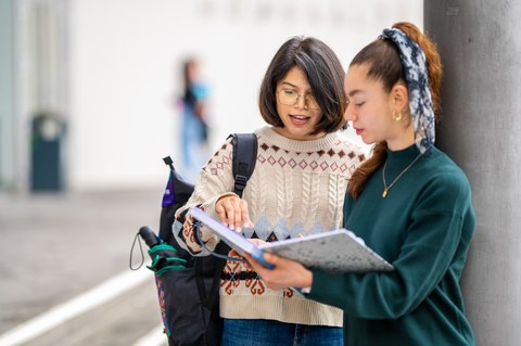 Foto zeigt, wie eine Studierende an einer Säule steht und ihre Notizen einer anderen Studierenden zeigt.