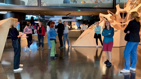 Ca. 10 Personen stehen in einer Ausstellungshalle und hören mit Kopfhörern einer Ausstellungsführerin zu. 