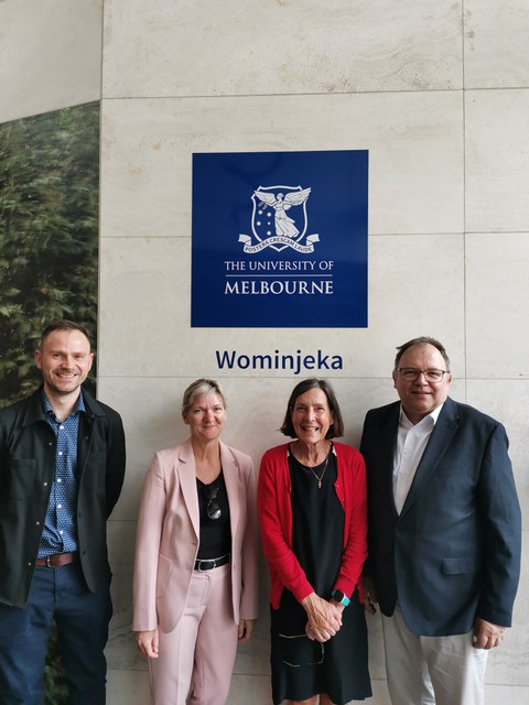 Vier Personen stehen vor einer Wand, auf der das Logo der Melbourne University zu sehen ist.