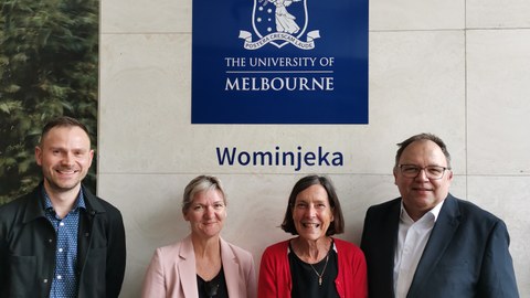 Vier Personen stehen vor einer Wand, auf der das Logo der Melbourne University zu sehen ist.