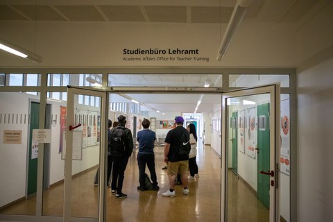 Eine Gruppe steht in einem Gang eines Unigebäudes. Über der Durchgangstür steht "Studienbüro Lehramt"