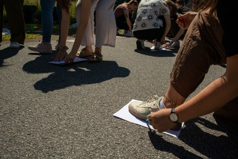 Zu sehen ist eine junge Frau, die im Vordergrund ihren eigenen Schuhabdruck zeichnet. Im Hintergrund sind weitere Personen zu sehen, die es ihr gleichtun.
