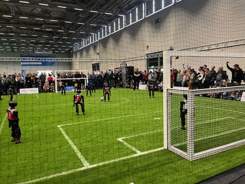 Zu sehen ist ein Fußballfeld in einer Messehalle, auf dem kleine Roboter gegeneinander Fußball spielen