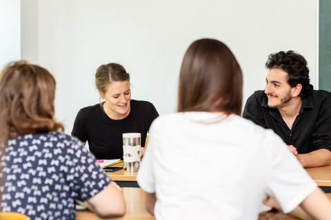 Foto von vier Personen, die sich an zwei Tischen gegenübersitzen und sich unterhalten.