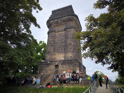 Das Foto zeigt den Bismarckturm in Dresden, vor dem Mitwirkende an der Gedenklesung sitzen und stehen.