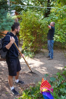 Mann mit Laubrechen. im HIntergrund schneidet ein weiterer Mann an einer Hecke