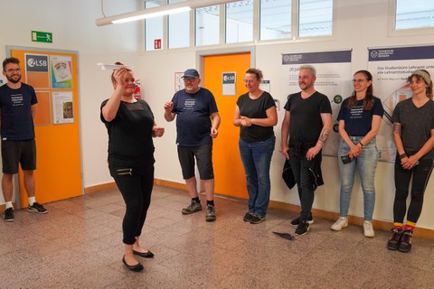 Frau steht mit Papierflieger in der Hand in einem Flur. Um sie herum stehen Leute, die den Wurf des Papierfliegers bestaunen.