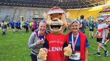 zwei Frauen im Sportoutfit stehen links und rechts neben einem Maskottchen
